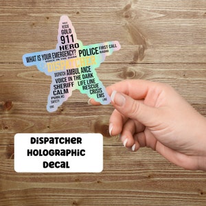 May include: A holographic decal in the shape of a star, featuring the word "DISPATCHER" and related terms like "POLICE," "911," and "RESCUE." The decal is held by a hand against a wooden background. The text "DISPATCHER HOLOGRAPHIC DECAL" is also present.