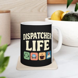 Puede incluir: Taza de cerámica blanca con la frase "DISPATCHER LIFE" en una fuente retro. Debajo del texto hay cuatro iconos: unos auriculares, un coche de policía, una taza de café y un marcador de mapa. La taza está sobre un posavasos beige.
