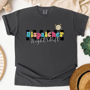 May include: A dark gray t-shirt with a graphic design featuring a cartoon bird and the text "dispatcher nightshift" in colorful retro letters.