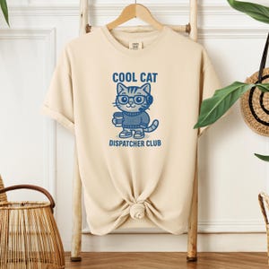 May include: Beige t-shirt with a blue graphic of a cat wearing glasses and a headset, holding a coffee cup. The text "COOL CAT DISPATCHER CLUB" is above and below the cat illustration.