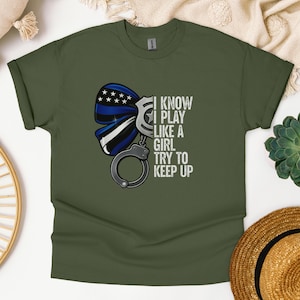 以下が含まれることがあります： オリーブグリーンのTシャツ。青と白のリボン、手錠、そして「I KNOW I PLAY LIKE A GIRL TRY TO KEEP UP」というテキストが特徴です。デザインは法執行機関をテーマにしています。