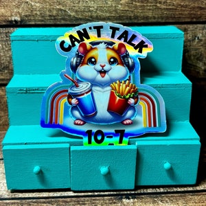 Pode incluir: Um adesivo hologr&aacute;fico com um hamster de desenho animado usando fones de ouvido e segurando um refrigerante e batatas fritas. O adesivo diz "Can't Talk 10-7" em uma fonte arco-&iacute;ris.