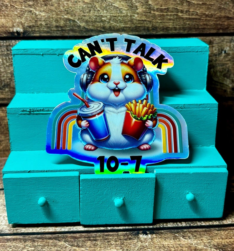 Pode incluir: Um adesivo hologr&aacute;fico com um hamster de desenho animado usando fones de ouvido e segurando um refrigerante e batatas fritas. O adesivo diz "Can't Talk 10-7" em uma fonte arco-&iacute;ris.