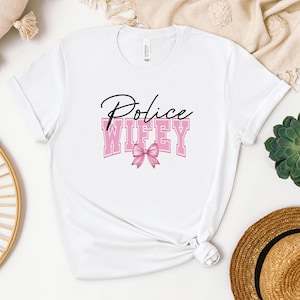 Peut inclure: T-shirt blanc avec le texte "Police Wife" en lettres roses avec un nœud rose.