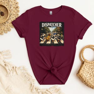 Puede incluir: Camiseta granate con un gráfico de cuatro patos cruzando una calle, parodiando la icónica portada del álbum Abbey Road. La palabra "DISPATCHER" está impresa encima de la imagen. La camiseta es de material suave.