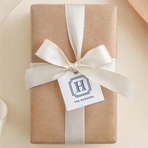 May include: A wrapped gift in brown paper, tied with a white satin ribbon and bow. A white tag with a blue framed "H" and the text "THE HUDSONS" is attached to the bow. The gift is presented on a light beige surface.
