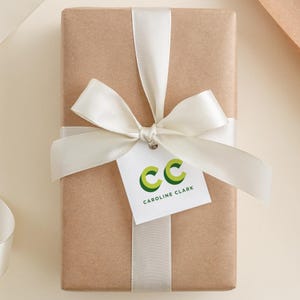 May include: A wrapped gift in brown paper, tied with a cream-colored ribbon and bow. A white tag with the initials "CC" in green and the name "Caroline Clark" hangs from the bow.