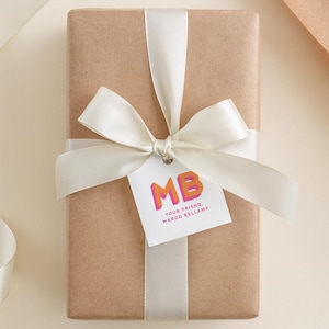 May include: A wrapped gift in brown paper, tied with a white satin ribbon and bow. A white tag with the letters "MB" in orange and pink, and the text "YOUR FRIEND, MARGO BELLAMY" is attached to the bow.