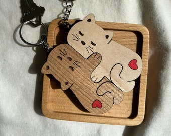 May include: A wooden keychain featuring two cats hugging each other. The cats are brown and white and have red hearts on their bellies. The keychain is attached to a silver key ring.
