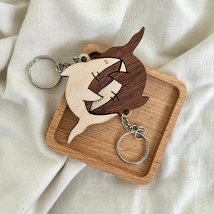 May include: A wooden keychain featuring two sharks, one brown and one natural wood, intertwined. The keychain is attached to a silver key ring.
