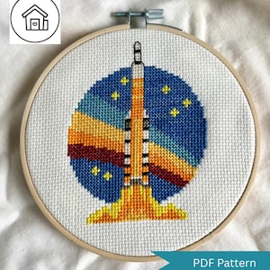 May include: A cross-stitch embroidery in a wooden hoop depicts a rocket launching into space. The design features a blue and yellow background with stars, and the rocket is orange and white. The words "PDF Pattern" are on a blue banner.