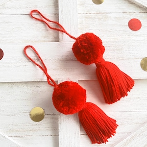 May include: Two red pom-pom ornaments with long red tassels hanging from white wooden boards.