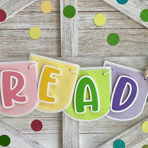 May include: A rainbow felt banner with the word "READ" in large, colorful letters. The banner is strung with wooden beads and colorful yarn tassels.