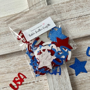 May include: A clear plastic bag filled with red, white, and blue star and letter-shaped confetti. The bag has a label that reads "Dearly Imagined table Scatter Confetti." The word "USA" is visible in red glitter. The confetti is scattered on a white wooden surface.