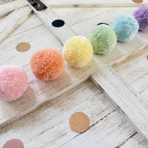 May include: A set of six colorful pom poms in a rainbow color scheme. The pom poms are arranged in a line on a white wooden surface.