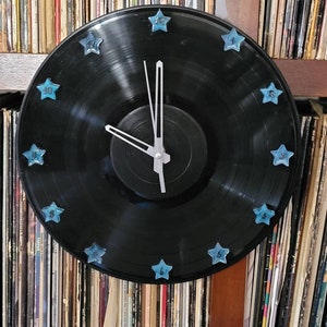 May include: A black vinyl record clock with blue star-shaped hour markers. The clock is hanging on a shelf with a collection of vinyl records behind it.