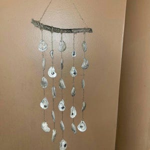 May include: A rustic oyster shell wind chime with a driftwood branch and natural twine. The shells are arranged in a cascading pattern, creating a whimsical and coastal-inspired decor piece.