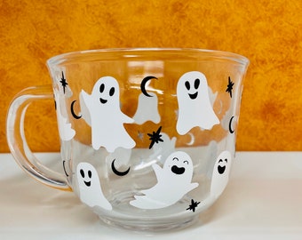 May include: A clear glass mug featuring a repeating pattern of white ghosts with black facial features, interspersed with small black crescent moons and four-pointed stars printed across the exterior surface of the round vessel.