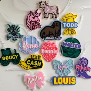 May include: A selection of vibrant luggage tags in diverse shapes, such as a cow, unicorn, otter, flower bouquet, seashell, strawberry, tractor, bulldozer, butterfly, bow, cowboy boot, and a round tag. Each tag has a name in a contrasting colour.