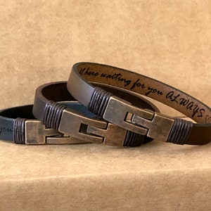 May include: Two leather bracelets with dark brown bands and antique bronze-toned metal clasps. The bracelets feature dark brown cord wrapping near the clasps. The interior of one bracelet is engraved with the text "here waiting for you Always".