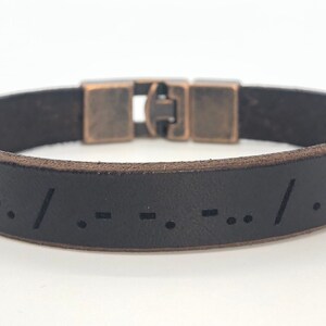 Puede incluir: Pulsera de cuero marrón oscuro con diseño de código Morse. La pulsera tiene un cierre de color bronce y mide aproximadamente 2 cm de ancho. El código Morse está grabado en el cuero.