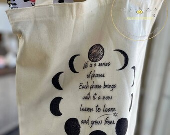 Peut inclure: Un sac fourre-tout en toile blanche avec un motif noir de cycle lunaire et le texte "La vie est une série de phases. Chaque phase apporte une nouvelle leçon à apprendre et à grandir."