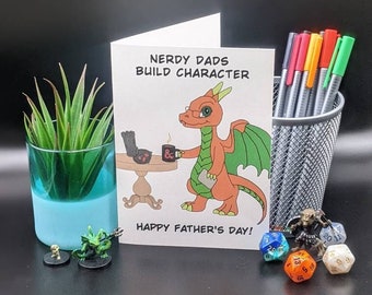May include: A Father's Day card with a cartoon dragon illustration. The card reads "NERDY DADS BUILD CHARACTER" and "HAPPY FATHER'S DAY!". The image also includes a small succulent, pens, and dice.