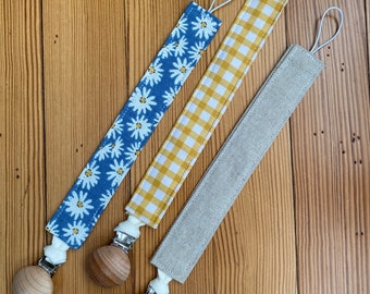 Könnte beinhalten: Drei Schnullerketten mit Holzperlen und verschiedenen Stoffmustern: Blau mit weißen Gänseblümchen, gelber Gingham und natürlicher Leinen.