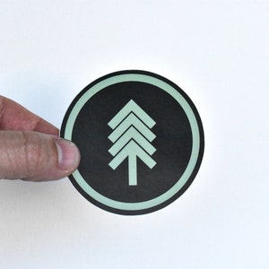 May include: A round sticker with a black background and a white outline. The sticker features a stylized green tree design.