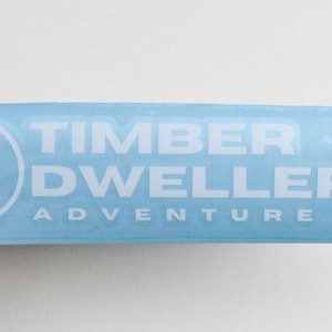 May include: White sticker with a blue background. The sticker features a logo with a stylized tree inside a circle and the text "TIMBER DWELLERS ADVENTURE CO."