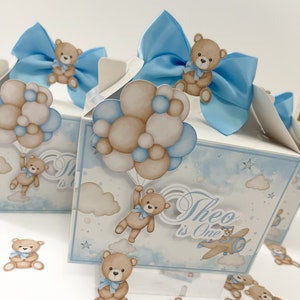 Puede incluir: Cajas de regalo azules y blancas con un osito de peluche sosteniendo globos, un osito de peluche volando en un avión y el texto "Theo is One". Las cajas están decoradas con nubes y estrellas. También hay pequeños recortes de ositos de peluche esparcidos alrededor de las cajas.