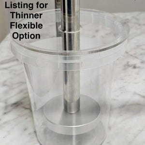 May include: A clear plastic cylindrical container with a metallic silver plunger mechanism inside. The text "See Other Listing for Thinner Flexible Option" is printed in black on the upper left side of the transparent container wall.