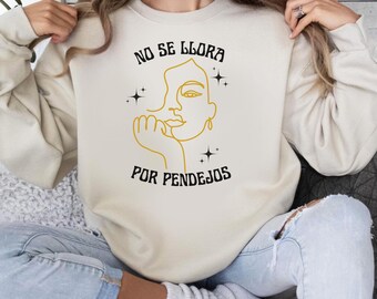 May include: Cream-colored sweatshirt with the text "NO SE LLORA POR PENDEJOS" in black, and a gold outline of a face. The design includes star accents. The sweatshirt is a casual style.