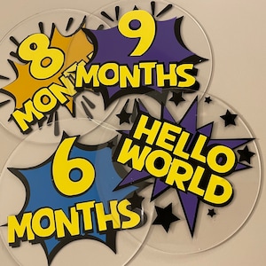 May include: Four round, clear acrylic milestone markers with colourful comic book style designs. The markers feature the numbers 6, 8, and 9 with the text "MONTHS" and one marker says "HELLO WORLD".