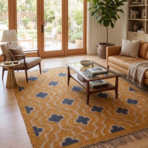 Puede incluir: Una alfombra rectangular con una base de color amarillo mostaza, un patrón geométrico blanco repetitivo y detalles en azul oscuro con forma de cuatrifolio. La alfombra tiene un tejido texturizado y flecos cortos de color marrón claro.