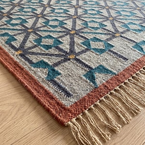Puede incluir: Una alfombra rectangular con una base gris claro y un patrón geométrico repetitivo en azul oscuro y verde azulado. El diseño incluye pequeños acentos amarillos en las intersecciones. La alfombra tiene un borde naranja quemado y flecos beige.