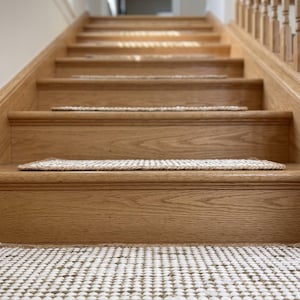 Op de afbeelding: Een set rechthoekige traptredenmatten gemaakt van een geweven mix van natuurlijke jute en witte vezels. Elke mat heeft een gestructureerd, rasterachtig patroon met een dikke, gevlochten jute rand, geïnstalleerd op houten traptreden.