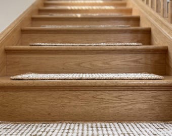 Könnte beinhalten: Ein Set rechteckiger Treppenstufenmatten aus einem gewebten Mix aus natürlicher Jute und weißen Fasern. Jede Matte weist ein strukturiertes, gitterartiges Muster mit einem dicken, geflochtenen Jutekantenabschluss auf den Holzstufen auf.