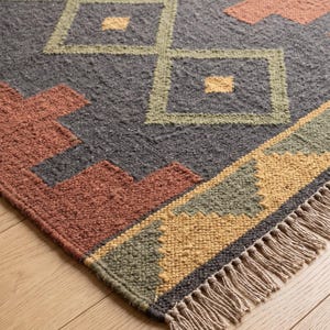 May include: A rectangular woven rug featuring a dark charcoal gray base with geometric patterns in terracotta, olive green, and mustard yellow. The design includes diamond shapes and stepped borders, finished with natural beige fringe on the ends.