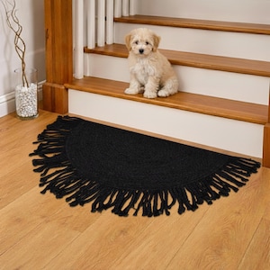 May include: A black, semi-circular rug with fringe detail sits at the base of a wooden staircase. A small, fluffy, light-colored dog sits on the stair. The rug is made of a woven material and adds a decorative touch to the entryway.