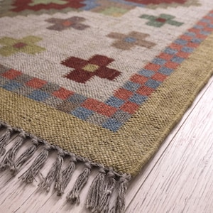 Può includere: Un tappeto intrecciato con una base color crema e un motivo ripetuto di piccole croci rosse, verdi e blu. Il tappeto presenta un ampio bordo verde oliva con un motivo a scacchi ed è rifinito con nappe a frangia di colore grigio.