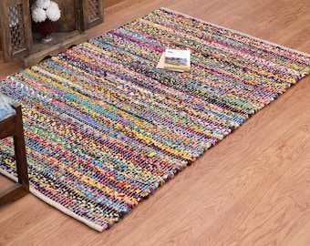 May include: A rectangular, multi-colored rag rug with a woven texture. The rug features a vibrant mix of colors, including pink, purple, blue, yellow, and green. A book rests on the rug. The rug is placed on a wooden floor.