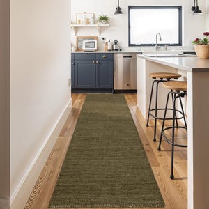 May include: An olive green runner rug in a kitchen. The rectangular rug is laid on a wooden floor. The kitchen has navy blue cabinets, stainless steel appliances, and a white countertop with wooden stools. The rug is approximately 300cm long.