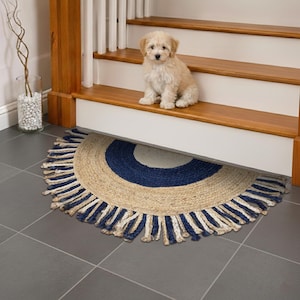 May include: A half-circle jute rug with a navy blue and cream design. The rug has a fringed edge and is placed on a gray tiled floor. A small, light-colored dog sits on the wooden stairs above the rug.