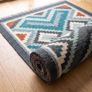 Puede incluir: Una alfombra rectangular con un patrón geométrico en tonos azul verdoso, azul claro, gris oscuro, crema y naranja quemado. La alfombra tiene un tejido grueso y texturizado, está parcialmente enrollada y muestra flecos en el borde.