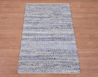 May include: A rectangular woven rug in shades of blue, grey, and beige. The rug has a textured appearance with horizontal stripes. The edges are finished with a light beige border. Suitable for a living room or bedroom.