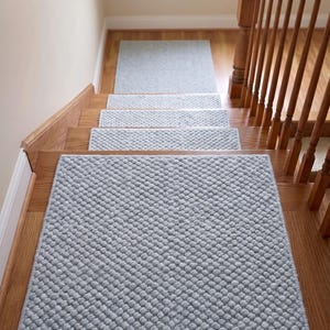 Peut inclure: Un ensemble de tapis d'escalier rectangulaires gris clair avec un motif en relief texturé rappelant des petites boules. Chaque tapis est placé au centre d'une marche en bois, créant un aspect uniforme sur tout l'escalier.