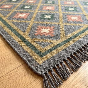 May include: A rectangular woven rug featuring a gray base with a yellow diamond grid pattern. Each diamond contains a small square motif in either muted pink or dark green with a white center. The rug has a dark green border and gray fringe.