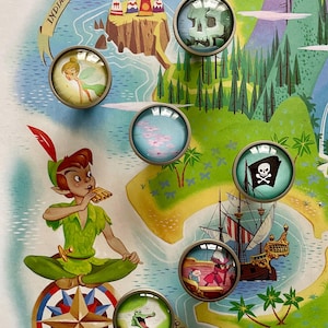 May include: A set of 8 round glass cabochons with various images from the movie Peter Pan. The images include Tinkerbell, a skull and crossbones, a compass, a crocodile, a pirate ship, a Native American village, and a pink sky. The cabochons are set in silver-toned metal settings.
