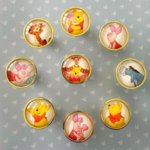 Peut inclure: Neuf boutons de meuble ronds avec des bords dorés. Chaque bouton présente une image différente des personnages de Winnie l'ourson, dont Winnie l'ourson, Tigrou, Porcinet et Bourriquet. Les boutons sont posés sur un fond bleu clair avec des icônes blanches de Mickey Mouse.